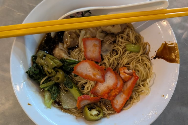A bowl of wonton noodles with char siu pork, leafy greens, and yellow chopsticks on a metal table.