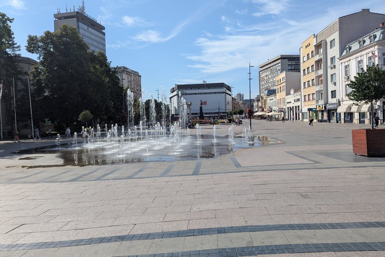 A wide stone plaza featuring a floor level water fountain and surrounding urban city buildings in NIs