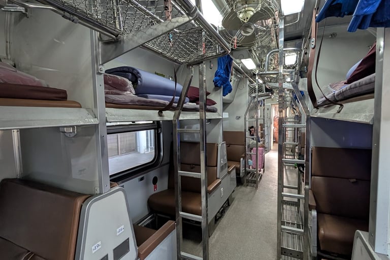 Interior of a Thai railway sleeper class coach featuring bunk beds, luggage racks, and ceiling fans.