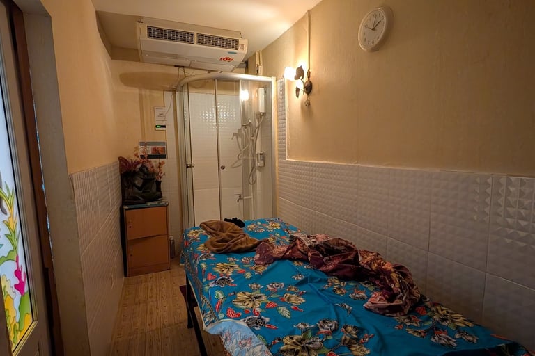 A narrow massage room with a floral blue bed, glass shower cubicle, and air conditioning unit.