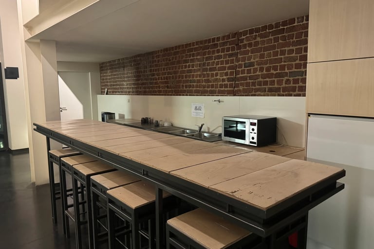 Cuisine de coworking avec longue table haute en bois, tabourets, évier, micro-ondes et mur en briques apparentes.