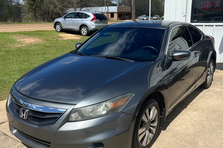 Dark gray Honda Accord coupe parked on a concrete lot at a used car dealership on a sunny day.