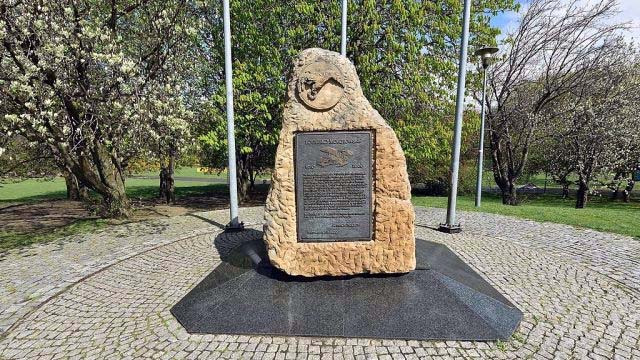 The obelisk for Mokotow Airport, in Warsaw, Poland. Sandy yellow coloured shaped rock with metal plaque inserted, in a park.