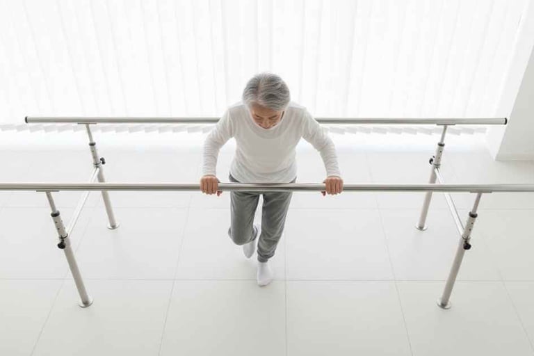 Vista superior de pessoa idosa realizando exercício de equilíbrio com apoio em barra em clínica de reabilitação.