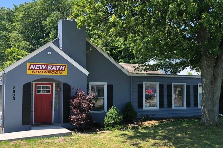 Front view of the New Bath Elite showroom in Perrysburg, Ohio, featuring a gray building with a red door, bright yellow logo 