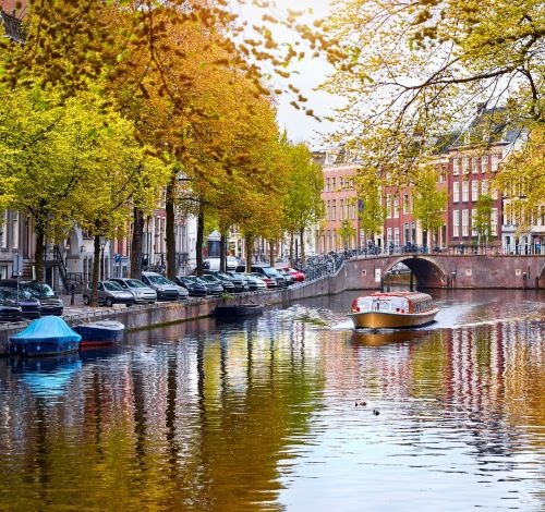 Tour boat cruising on an Amsterdam canal lined with historic buildings and autumn trees.