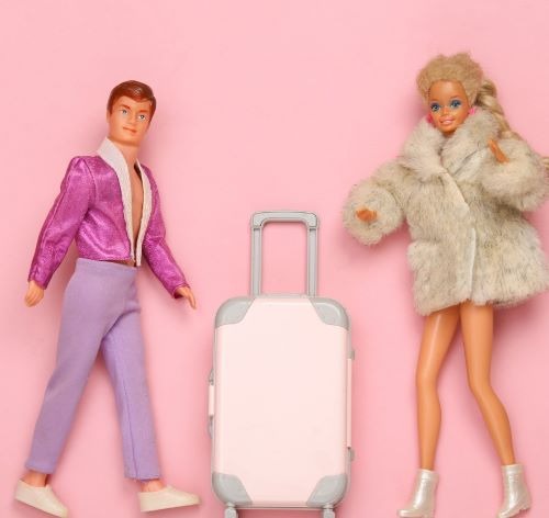 Ken and Barbie dolls in stylish outfits posing with a pink toy suitcase on a pink background.
