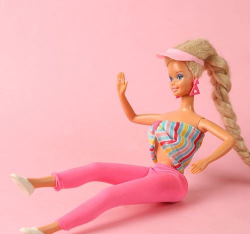 Blonde Barbie doll in 80s workout clothes with pink leggings and striped top against a pink background.