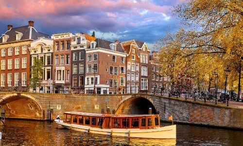Historic Amsterdam canal houses with a tourist boat cruising under a stone bridge at sunset.