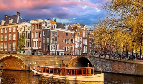 Tour boat cruising on an Amsterdam canal past historic canal houses under a pink sunset sky.