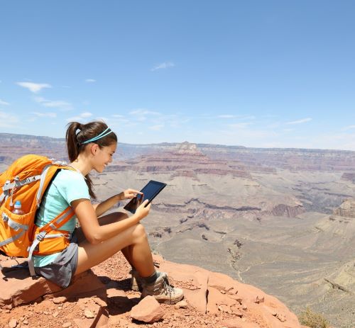 Female hiker with orange backpack sitting on a canyon rim using a digital tablet.
