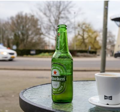 A green Heineken beer bottle and a cup of coffee on an outdoor glass table by a street.
