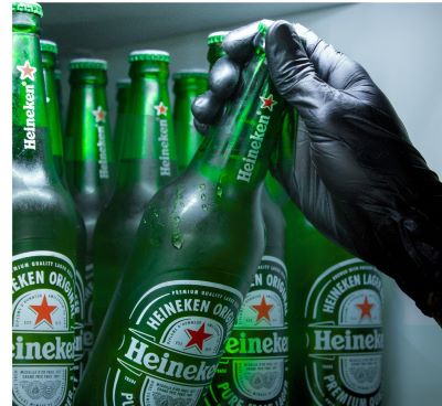 A gloved hand grabbing a cold bottle of Heineken lager beer from a refrigerator shelf.