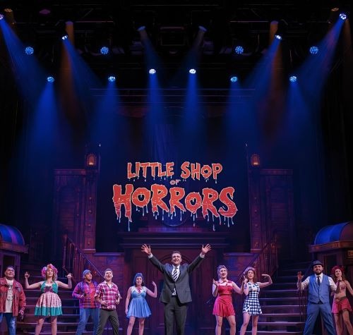 a man in a suit and tie standing on stage with the sign “little horrors”
