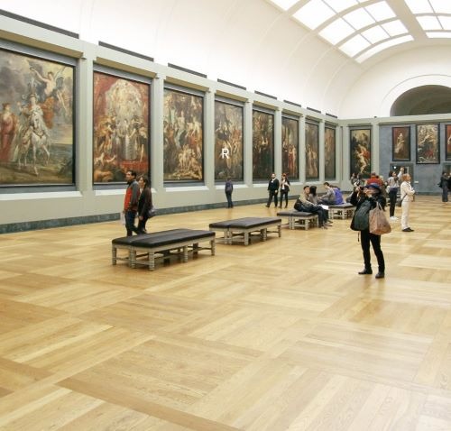 Visitors view large classical oil paintings in a bright art museum gallery with parquet floors.
