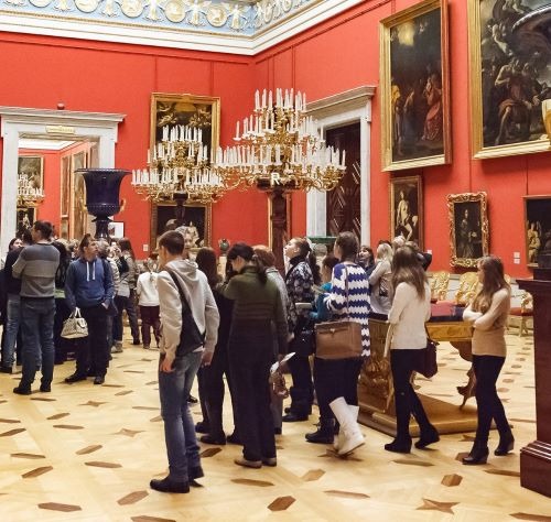 Tourists viewing classic oil paintings in an ornate red art gallery with chandeliers.