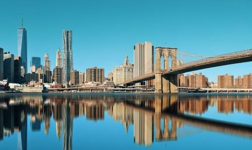 Panoramic Lower Manhattan skyline and Brooklyn Bridge reflecting in the East River under a clear blue sky.