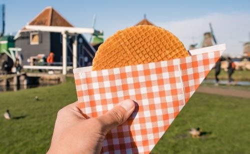 Hand holding a Dutch stroopwafel in a checkered sleeve with Zaanse Schans windmills in the background.