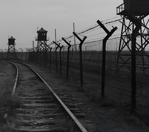 Black and white image of railway tracks leading past a barbed wire fence and watchtowers.