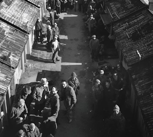 A high-angle black and white historical photo of people walking through a narrow street of wooden shelters.