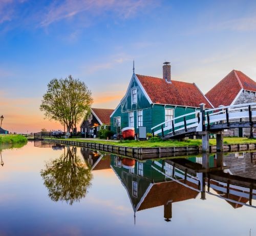 Traditional green Dutch houses reflecting in a calm canal at sunset in Zaanse Schans village.
