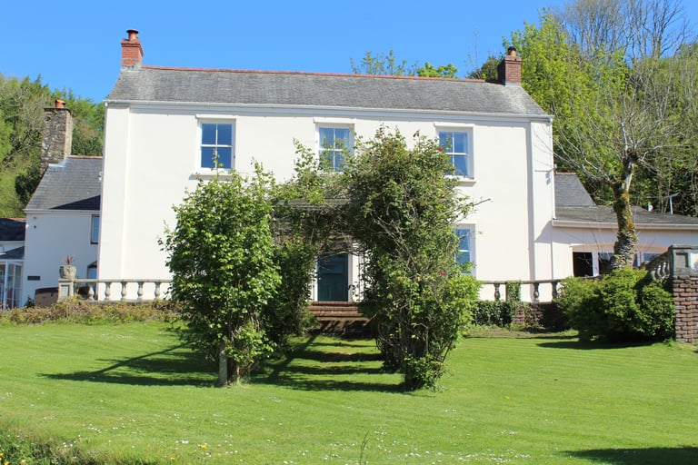 A white two-story country house with a slate roof and green garden hedges.