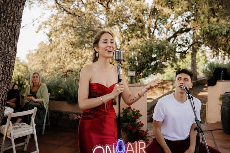 A female singer in a red dress performing with a vintage microphone at an outdoor garden wedding event.