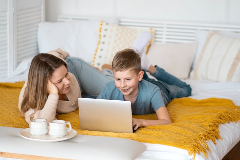 Smiling mother and son lying on a clean white bed, watching something together on a laptop
