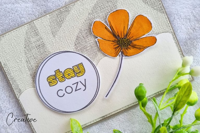 Handmade greeting card with "Stay cozy" sentiment and big orange flower on a beige background.