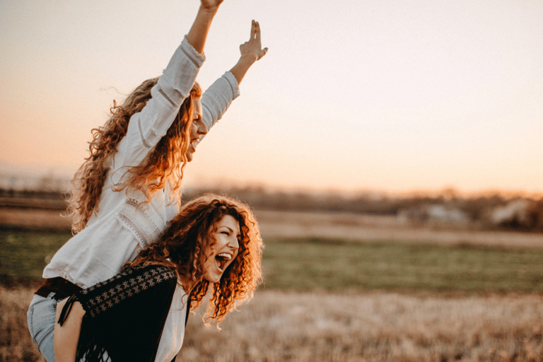 two joyful women with curly long  hair are celebrating, one lifting the other up on her shoulders 