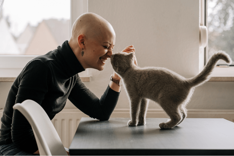 a smiling woman in a black turtle neck and a  bald head and a grey cat are touching nose to nose