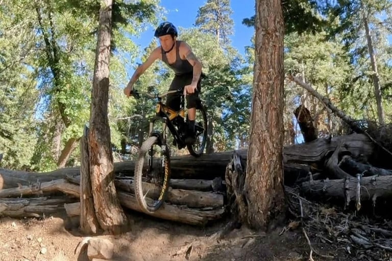 Mark Hower riding a mountain bike over a fallen log in a pine forest in Southern California, black