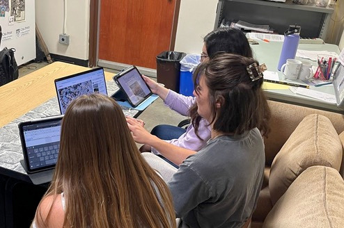 Three students sitting at a table, each with a computer or tablet in front of them reviewing video