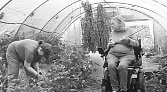 a black and white photo of two people in the greenhouse. One person sits in a motorized wheelchair