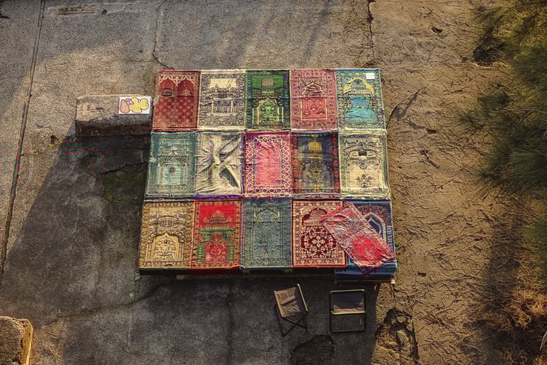 An aerial view of 16 prayer rugs arranged in a grid in an outdoor concrete setting.