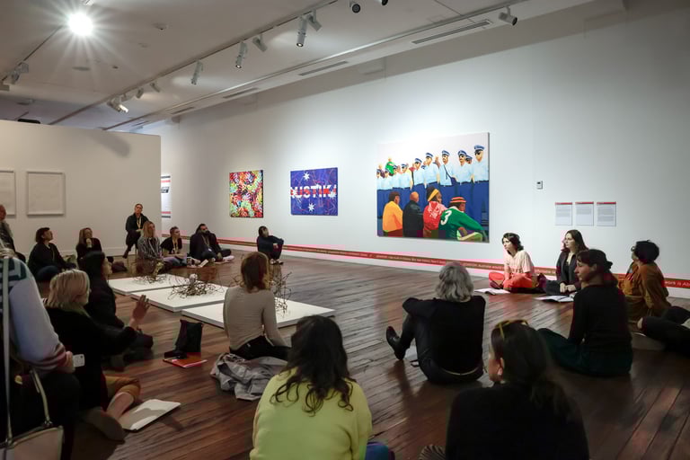 A group of people sitting in a loose circle discussing paintings by Richard Bell.