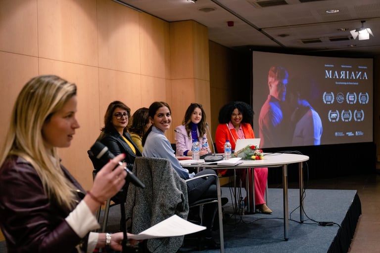 A panel of female speakers sits at a table during a film presentation for Mariana in an auditorium.