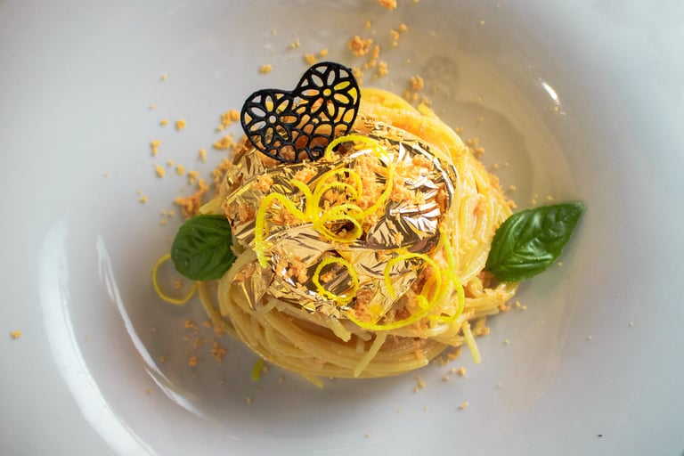 Spaghetti con foglia d'oro, cuore nero traforato, briciole arancioni e basilico in piatto bianco