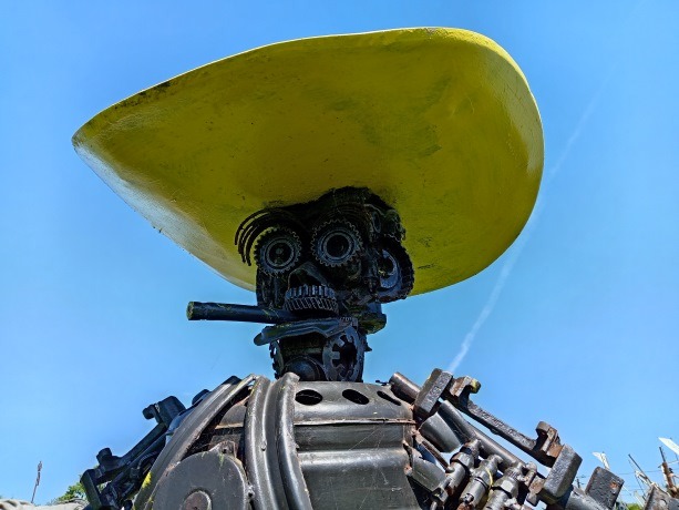 A metal scrap sculpture of a figure wearing a large yellow hat against a clear blue sky.