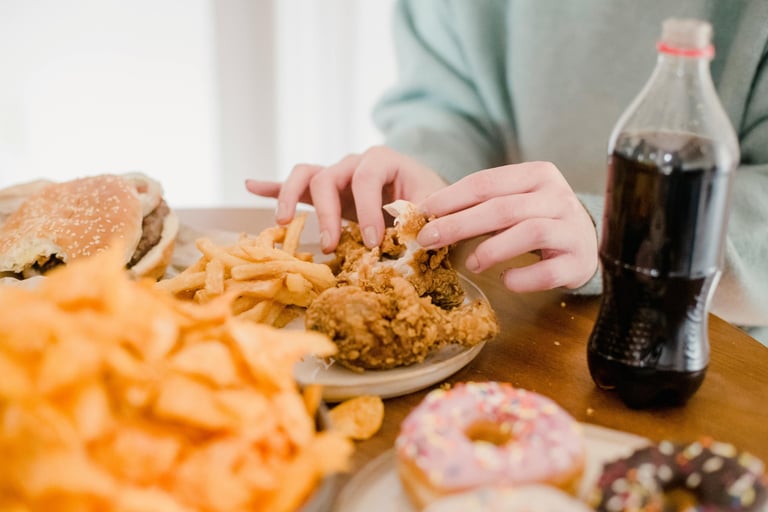 Person eating fried chicken with a side of french fries, burgers, donuts, and a cola soda.