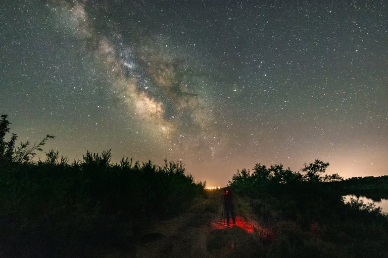 A photographer stands under the bright Milky Way galaxy in a starry night sky above a dark landscape.