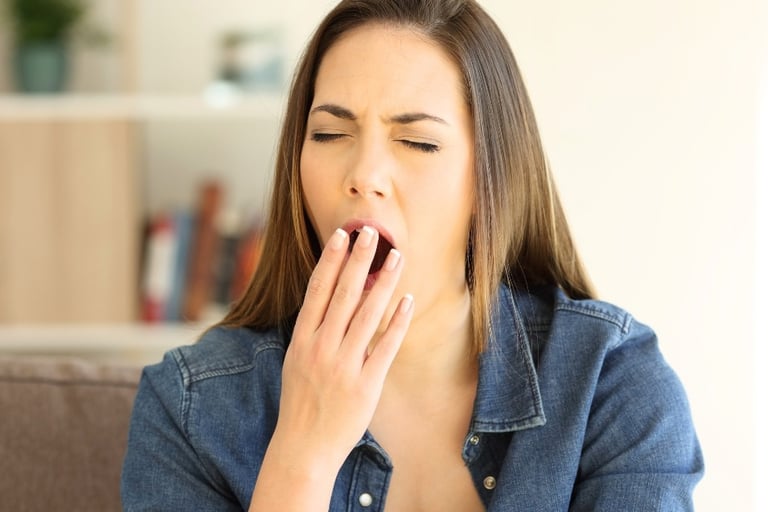 A tired young woman yawning and covering her mouth with her hand while sitting on a sofa.