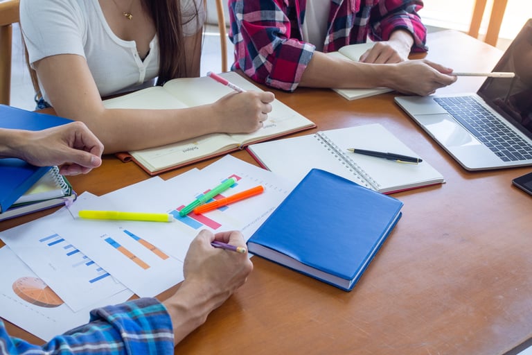 a group of people studying at a table with notebooks, pens, papers and a laptop