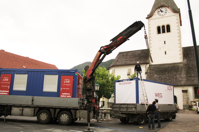 Kran bei der Aufstellung des Ausstellungscontainers "Aus weg los!" auf einem Dorfplatz.