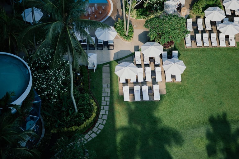 Aerial view of a tropical resort garden with white patio umbrellas and lounge chairs on a lush green lawn.