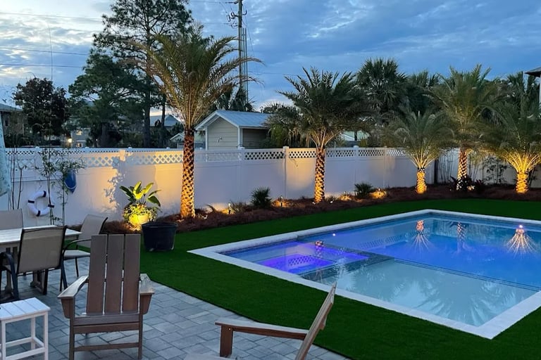 Backyard swimming pool with artificial turf and palm trees illuminated by warm landscape lighting.