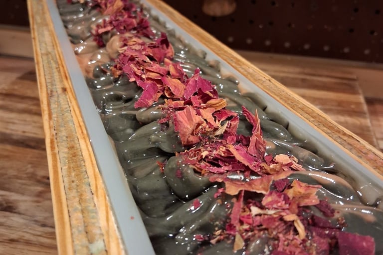 Handmade artisan loaf of dark charcoal soap topped with dried rose petals in a wooden mold.