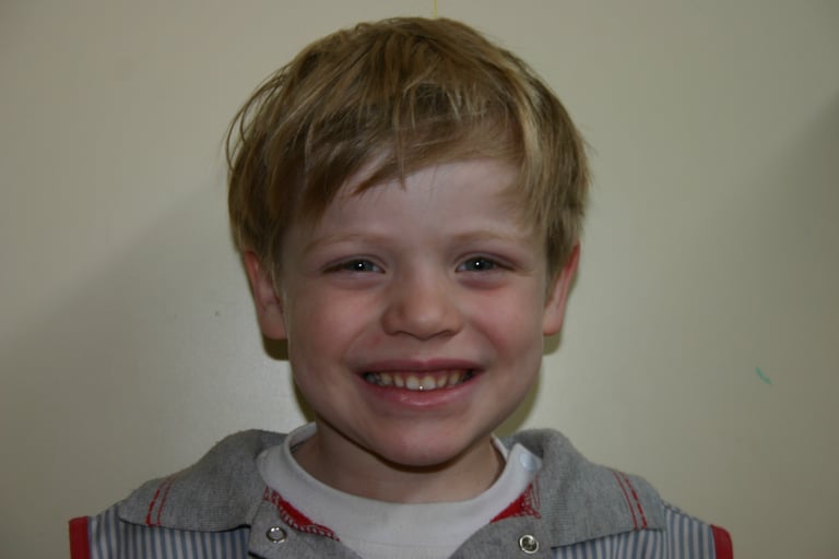 Smiling young boy with messy blonde hair posing for a portrait against a plain wall.