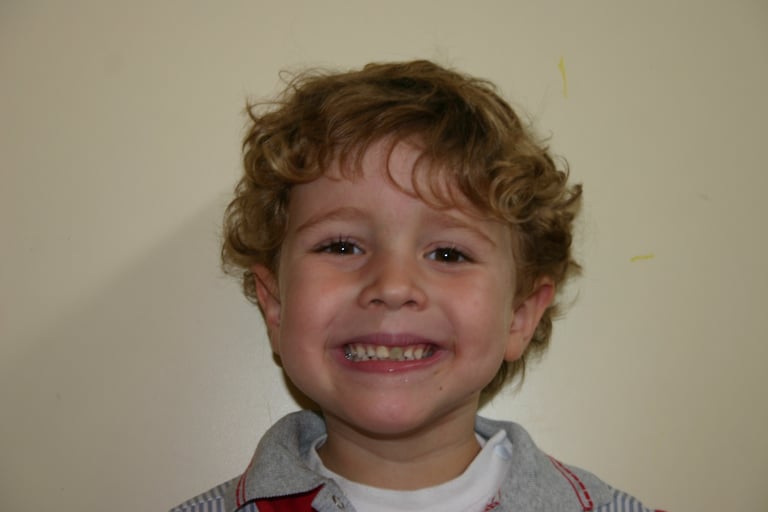 A smiling young boy with curly blonde hair showing his teeth in a happy portrait.
