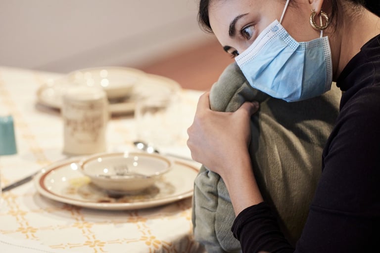A woman wearing a mask holds a vibrating cushion filled with chai spices at a kitchen table 